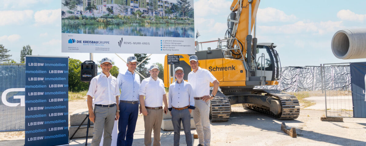 5 Projektbeteiligte stehen vor einem großen Plakat mit einem Gebäude drauf daneben steht ein Bagger